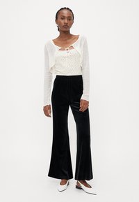 Witte geribbelde cropped cardigan met lange mouwen over een witte top met strikdetail. Zwarte fluwelen wijde broek en witte hakken.