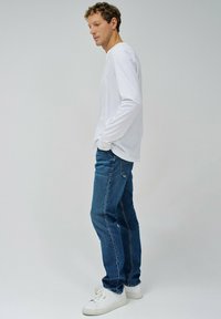 Weißes Langarmhemd, blaue Jeans und weiße Turnschuhe. Die Jeans haben eine klassische Passform, und das Hemd hat einen Rundhalsausschnitt.