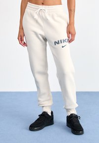 Calças de fato de treino cor creme da Nike com cintura elástica e punhos elásticos, apresentando letras e logotipo "NIKE" em azul marinho na coxa esquerda.