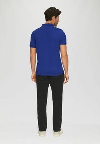 Herren Polo-Shirt in Blau mit kurzen Ärmeln, aus einem weichen Stoff gefertigt, kombiniert mit schwarzen Hosen und hellen Sneakers. Ansicht von hinten.