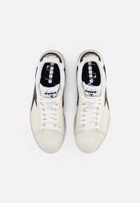 White leather sneakers with black accents, circular perforations, and white laces. Features a black insole with a logo. Flat rubber sole.