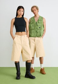 Top corto nero, pantaloni beige a gamba larga, gilet verde con più tasche. Stivali neri al ginocchio, sneakers marroni, su uno sfondo verde erba.