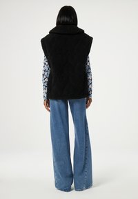 Zwarte, gewatteerde mouwloze vest met een hoge kraag, gedragen over een bloemenblouse met lange mouwen, gecombineerd met loszittende, lichtblauwe jeans.