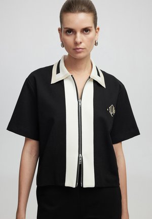 Chemise noire à manches courtes avec une fermeture éclaire blanche à l'avant, présentant un col contrasté et des rayures verticales. Détail logo doré sur la poitrine.