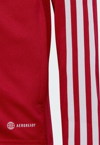 Rotes Sporttrikot aus strukturiertem Stoff. Verfügt über drei weiße Streifen, die entlang des Ärmels verlaufen, sowie ein "AEROREADY"-Logo am Saum.