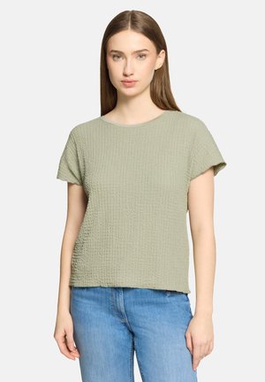 Junge Frau mit glatten braunen Haaren, die ein strukturiertes hellgrünes kurzärmeliges Oberteil und blaue Jeans trägt, vor einfachem Hintergrund stehend.