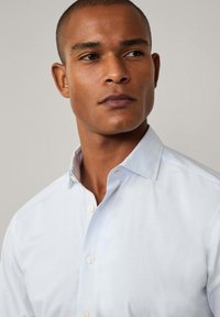 Light blue button-up shirt with a subtle check pattern, large collar, and white buttons. Smooth fabric with a slightly tailored fit.