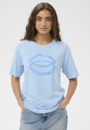 Kobieta o kręconych włosach ubrana w jasnoniebieski t-shirt z teksturowanym wzorem ust, w połączeniu z niebieskimi jeansami, ręce w kieszeniach.