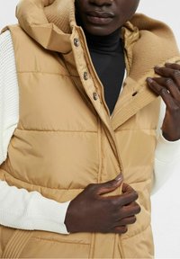 Gilet imbottito color tan con cappuccio, caratterizzato da bottoni a pressione e colletto a coste. Texture leggermente trapuntata e rivestimento interno beige a contrasto.