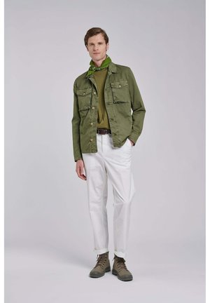Mężczyzna stoi, ubrany w oliwkową kurtkę, pasujący sweter, zieloną chustę na szyi, białe spodnie, brązowe buty, neutralne tło.