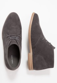 Chaussures chukka en daim gris avec deux œillets, semelle en caoutchouc neutre et détails de couture contrastants. Texture douce, design minimaliste.