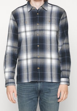 Mladá osoba stojící na jednobarevném pozadí, oblečená v modré, bílé a šedé kárované košili na knoflíky s dlouhým rukávem a modrých džínách.