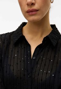 Blusa nera trasparente con righe testurizzate, paillettes e colletto. Presenta una chiusura a bottoni e dettagli di cucitura visibili.