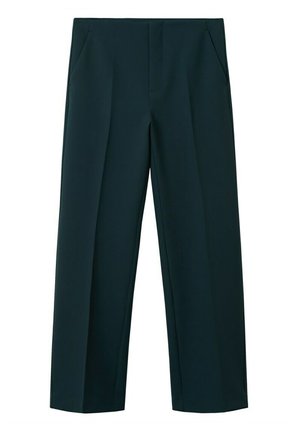 Frau trägt beigefarbenen Trenchcoat, weißen kurz geschnittenen Rollkragenpullover, dunkelgrüne weite Hosen und schwarze Schuhe mit Blockabsatz.