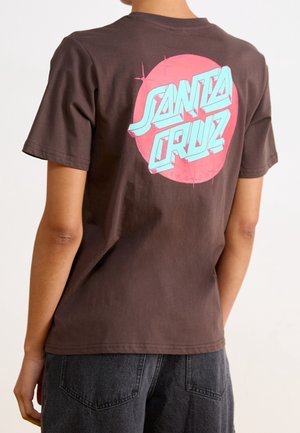 Osoba nosząca brązowy t-shirt z dużym różowym kołem i niebieskim napisem „Santa Cruz” na plecach.