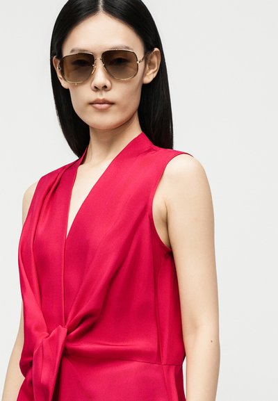 Femme aux longs cheveux noirs portant des lunettes de soleil marron et un top rose profond sans manches avec un nœud sur le devant, sur un fond uni.