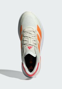 Zapatilla de running en color crema con rayas naranjas y acentos rosas. Parte superior de malla con detalles texturizados. Suela acolchada para mayor comodidad.