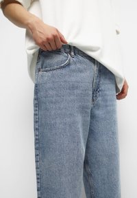 Persona vestida con jeans de mezclilla azul claro y una camiseta de manga larga blanca, con una mano descansando cerca del bolsillo delantero de los jeans.
