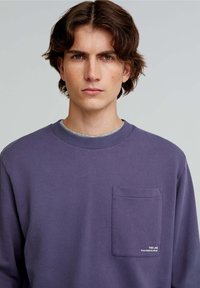 Paarse katoenen sweatshirt met een ronde hals, voorzien van een voorzak en geborduurd merklogo aan de onderkant. Soepele textuur, casual ontwerp.
