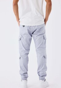 Uomo in pantaloni cargo grigi chiari e sneakers bianche, in piedi con le mani nelle tasche su uno sfondo neutro, vista posteriore.