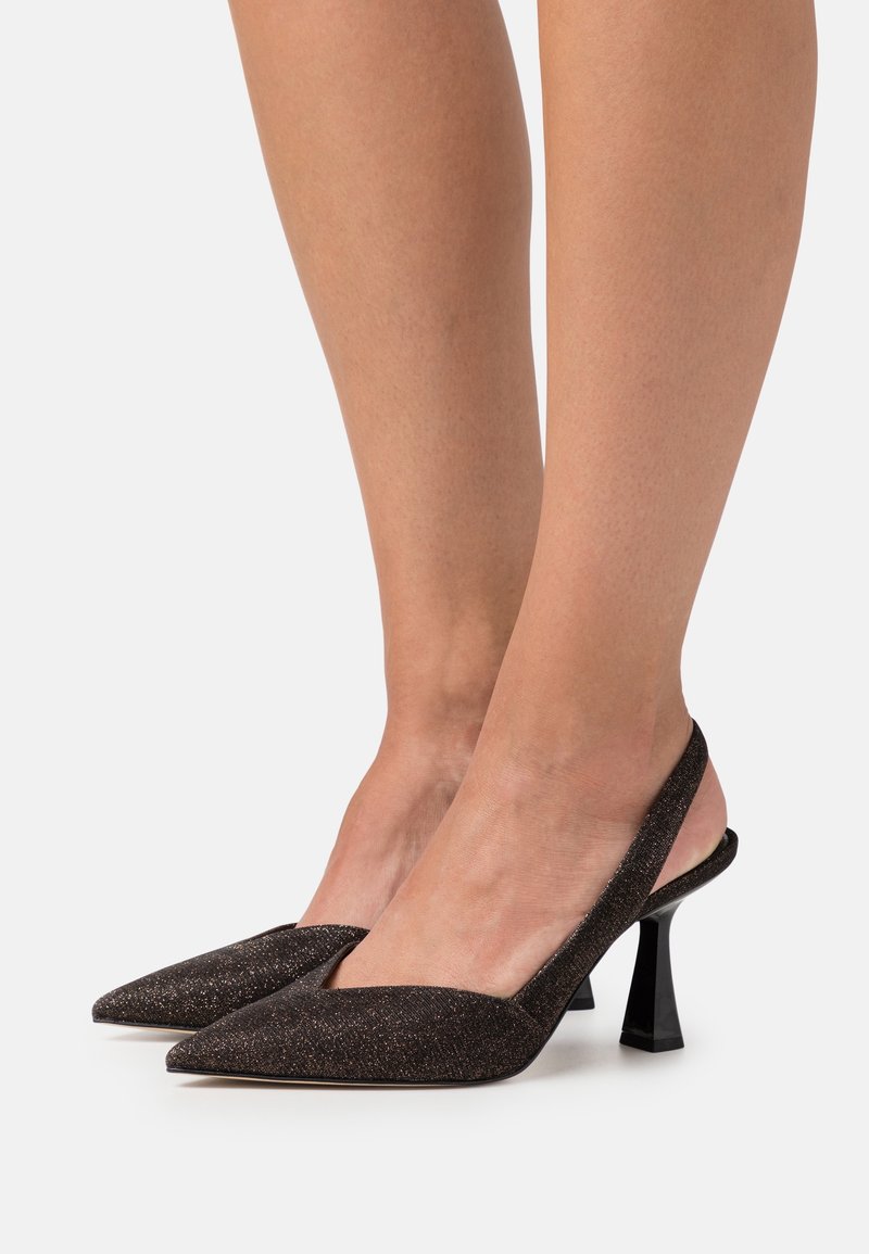 Kobieta w czarnych, błyszczących butach na obcasie typu slingback z odkrytymi palcami i słupkowym obcasem, stojąca na białym tle.