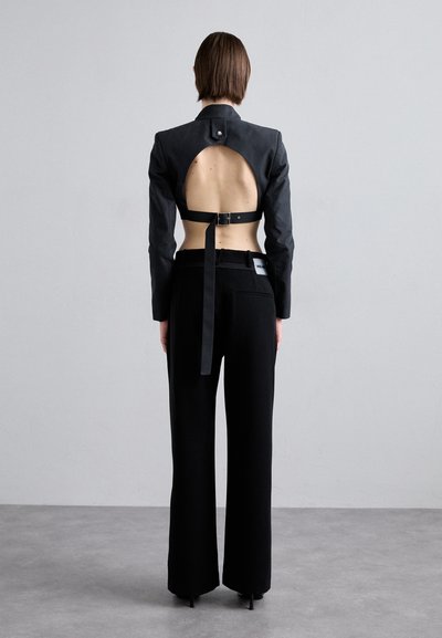 Black structured jacket with open back design, featuring a belt and button detail. Paired with wide-leg black trousers. Smooth fabric texture.