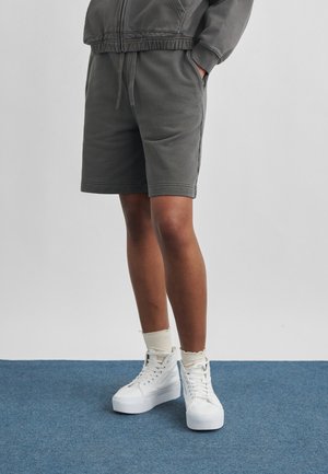 Persona vistiendo pantalones cortos informales gris oscuro, zapatillas blancas de caña alta y calcetines blancos, de pie sobre una alfombra azul contra un fondo claro y liso.