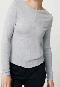 Mujer vestida con una camiseta ajustada de manga larga en gris claro con una costura vertical y pantalones negros, contra un fondo liso.