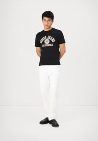 Camiseta de algodón negra con impresión en blanco y beige de "Guess Jeans California", combinada con pantalones de denim blancos y zapatos slip-on negros.