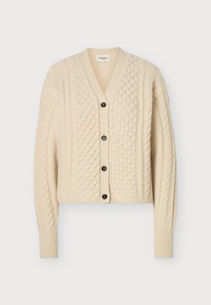 Femme portant un cardigan beige en maille torsadée avec des boutons noirs, une chemise blanche, un pantalon noir ample et des chaussures noires brillantes, debout devant un fond uni.