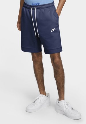 Mężczyzna ubrany w granatowe spodenki Nike z sznurkiem i białe buty Nike, stojący z rękami w kieszeniach na jednolitym tle.