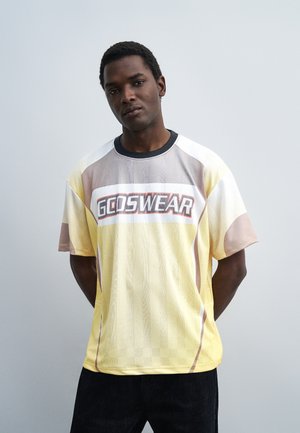 Άνδρας με σκουρόχρωμο δέρμα που φορά κίτρινο και μπεζ κοντομάνικο πουκάμισο GODSWEAR στέκεται μπροστά σε απλό ανοιχτό γκρι φόντο.