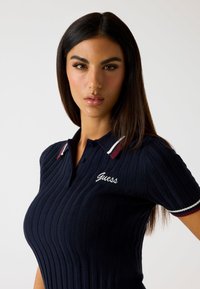 Polo shirt navy a coste con maniche corte, caratterizzato da accenti strisciati bianchi e bordeaux sul colletto e logo "Guess" ricamato.