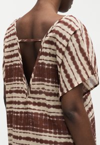 Άτομο που φοράει ένα καφέ και μπεζ ριγέ τοπ με κοντά μανίκια, βαθύ V στην πλάτη και οριζόντιο λουράκι στο πάνω μέρος της πλάτης.