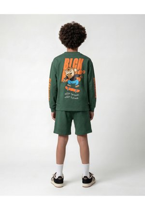 Kind dat groene shorts en een shirt met lange mouwen draagt met een cartoon-aap die skateboart en de tekst "KEEP TRYING. KEEP FLYING." op de rug, staand met de rug naar ons toe.