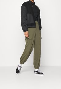 Veste bomber noire à texture matelassée, associée à un pantalon cargo vert olive avec un ourlet élastique. Des baskets noires et blanches complètent le look.