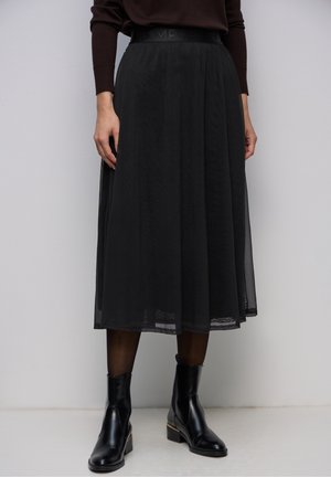 Jupe midi noire avec superposition transparente portée avec des bottines noires et des collants foncés sur un fond clair uni.