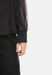 Schwarzes Oberteil mit transparenten langen Ärmeln und weißen Polka-Dots sowie elastischen Bündchen; kombiniert mit einem strukturierten schwarzen Unterteil.