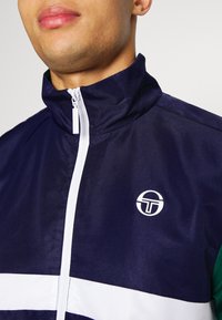 Námořnická bunda s bílým zipem a logem na přední straně. Obsahuje design s barevnými bloky s bílými a zelenými akcenty. Texturovaná tkanina se stojacím límcem.