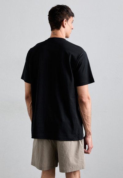 Μαύρο βαμβακερό T-shirt με κοντά μανίκια, στρογγυλή λαιμόκοψη και χαλαρή εφαρμογή. Φορεμένο με καρό σορτς, απεικονίζεται από πίσω.