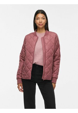 Femme portant une veste matelassée mauve à fermeture éclair sur un pull rose clair et un pantalon noir, debout devant un fond blanc uni.
