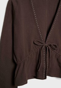 Rjava bluzica iz teksturirane tkanine z dolgimi rokavi, V-izrezom obrobljenim z majhnimi srebrnimi zakovicami, sprednjo pentljo in peplum gornjim delom.