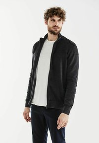 Giacca zip-up grigia scura con dettagli a coste sulle spalle, abbinata a una maglietta bianca. Il tessuto sembra morbido e texturizzato.