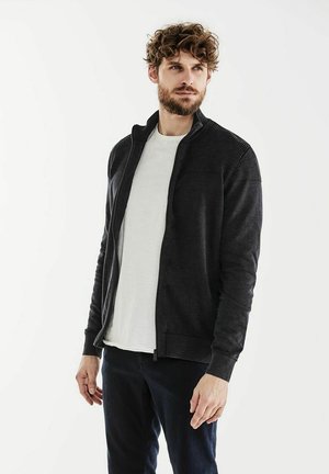 Giacca zip-up grigia scura con dettagli a coste sulle spalle, abbinata a una maglietta bianca. Il tessuto sembra morbido e texturizzato.