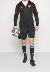 Personne portant un haut Manchester United noir, un short noir, ainsi que des chaussettes et chaussures Adidas, tenant un ballon de football de la Ligue des champions de l'UEFA.