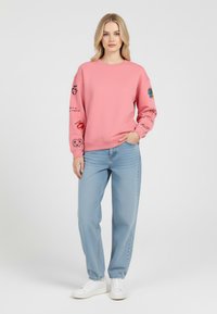 Felpa rosa con stampe grafiche sulle maniche, scollatura rotonda e polsini a costine. Abbinata a jeans larghi di colore blu chiaro e sneakers bianche.