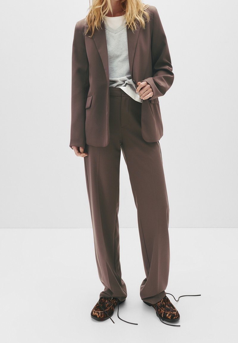 Personne portant un costume marron avec un pantalon à jambes larges et un blazer, un pull gris en dessous, et des chaussures imprimé léopard avec des lacets lâches sur fond blanc.
