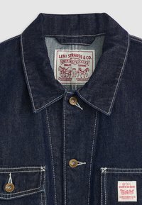 Denimjakke i mørkeblått med krage, to frontlommer og metallknapper. Har en sydd etikett med rød og hvit tekst.