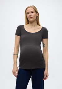 Trudnica sa plavom kosom, odjevena u usku tamno sivu majicu kratkih rukava i tamnoplave hlače, stoji ispred jednostavne bijele pozadine.