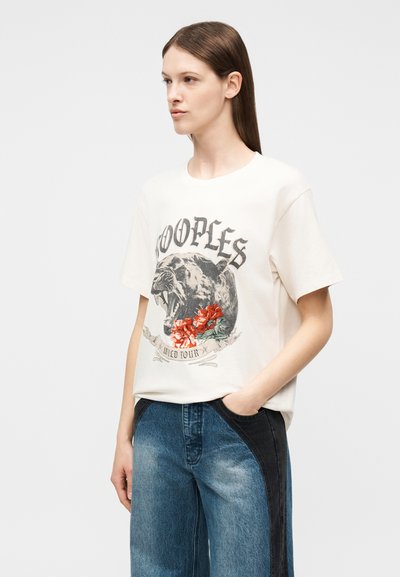 Γυναίκα με μακριά καστανά μαλλιά που φοράει κρεμ μπλουζάκι με ένα σχέδιο από μια βρυχώμενη πάνθηρα και κόκκινα λουλούδια, καθώς και τζιν παντελόνια διχρωμίας μπλε και μαύρου.